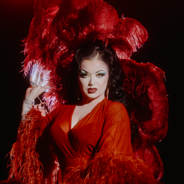 Performer in a red velvet dress hold a large red feather fan behind their head