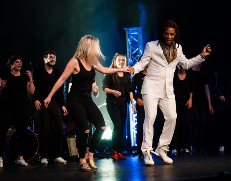Woman wearing black and a man wearing a white suit tap dancing together while holding hands