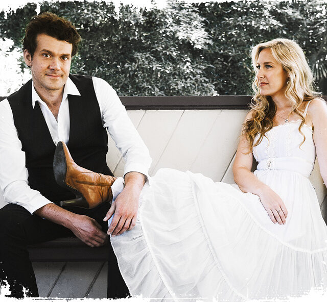 Members of The Weeping Willows Laura Coates in a white dress and Andrew Wigglesworth in a white shirt and black vest sitting on a bench