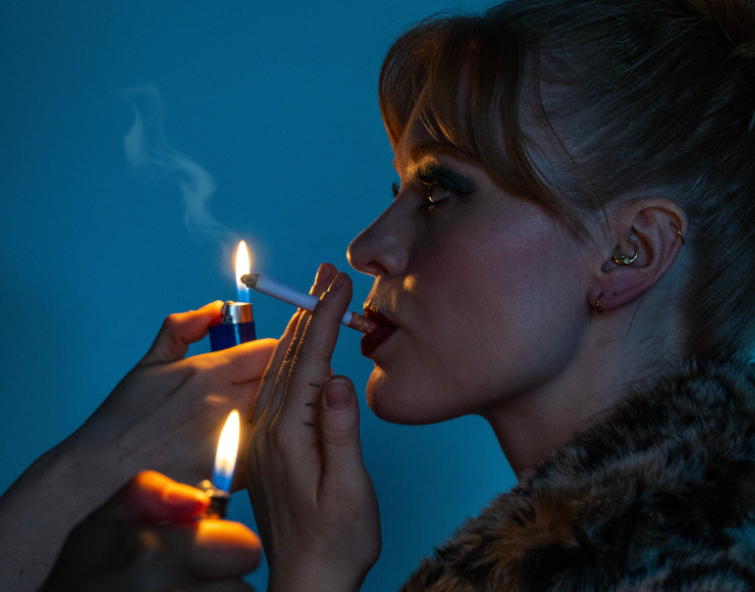 Profile head shot of a person with a cigarette in their mouth and two lighters