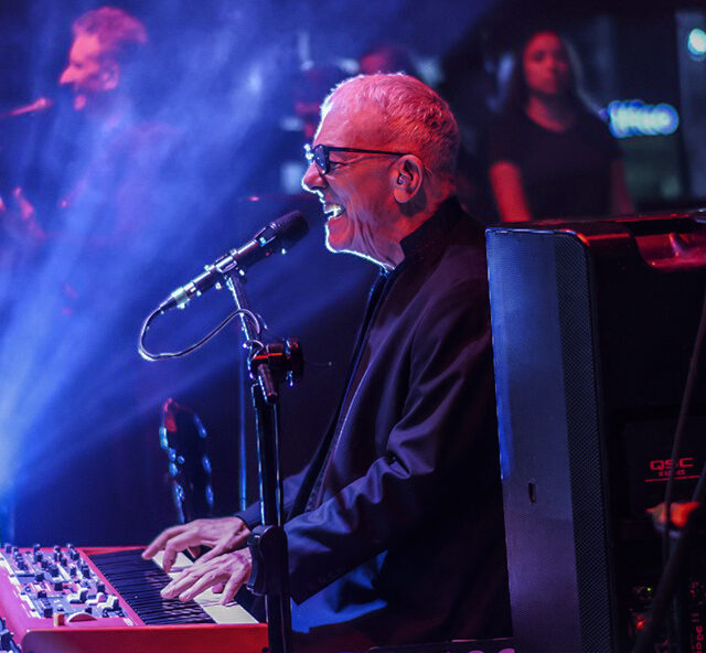 Musician Ilan Chester performs passionately at a keyboard on a dimly lit stage, singing into a microphone as blue and purple lights create a dramatic concert atmosphere.