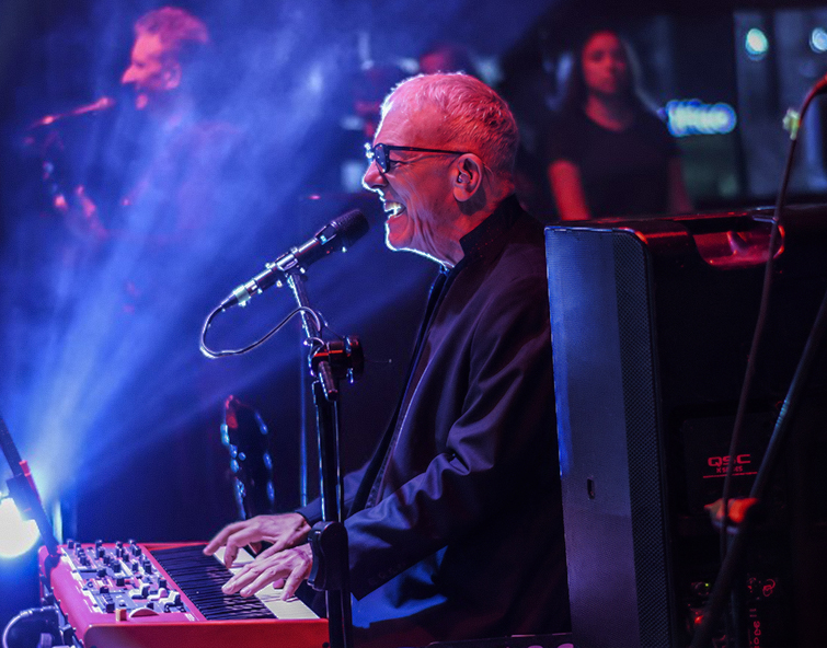 Musician Ilan Chester performs passionately at a keyboard on a dimly lit stage, singing into a microphone as blue and purple lights create a dramatic concert atmosphere.