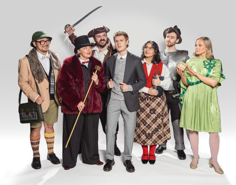 Seven adults pose together in eccentric, theatrical costumes against a plain white background. Their outfits include: • A man dressed as an explorer wearing a bucket hat and a tan jacket, a woman wearing a large burgundy faux-fur coat over a striped suit, a man standing in a pirate costume, a man wearing a modern grey blazer, grey trousers, and a knitted sweater, a woman wearing glasses, a white collared shirt under a red vest, and a long plaid skirt, a man dressed in silver medieval knight armor with a dark hat, and a woman on the far right wearing a bright green vintage-style dress with a matching green bow, holding a small decorative box.