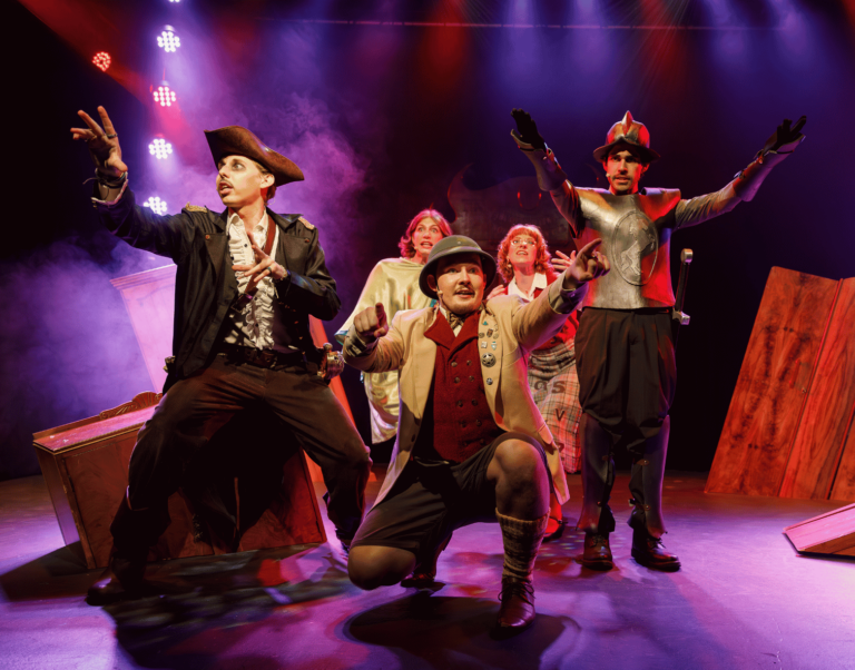 A theatrical group performs in colourful costumes on a stage, illuminated by dramatic purple and red lighting and surrounded by stage props and haze.
