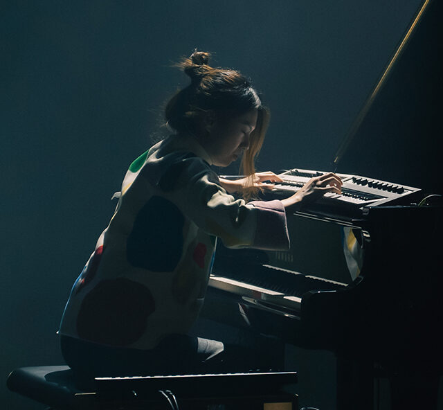 Belle Chen sits at a piano in a dimly lit room, playing the keys with focused concentration. Dramatic lighting highlights the performer and piano against a dark background.