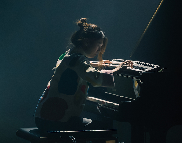 Belle Chen sits at a piano in a dimly lit room, playing the keys with focused concentration. Dramatic lighting highlights the performer and piano against a dark background.