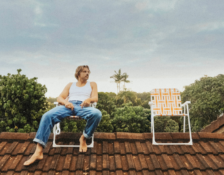 Artist Kylie Lionhart wears a white tank top and jeans sitting barefoot on a chair atop a tiled roof, with a colourful empty chair beside him and lush tropical trees under a cloudy sky.