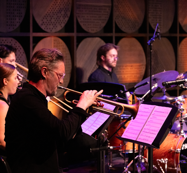 A jazz ensemble performs on stage, with a trumpet player in the foreground and other musicians playing trombone, piano and drums behind him. Warm orange and purple lighting creates an intimate concert atmosphere.