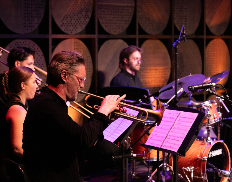 A jazz ensemble performs on stage, with a trumpet player in the foreground and other musicians playing trombone, piano and drums behind him. Warm orange and purple lighting creates an intimate concert atmosphere.