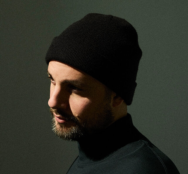 A moody portrait of a bearded Shai Maestro wearing a black beanie and dark turtleneck, lit from the side against a dark green background.