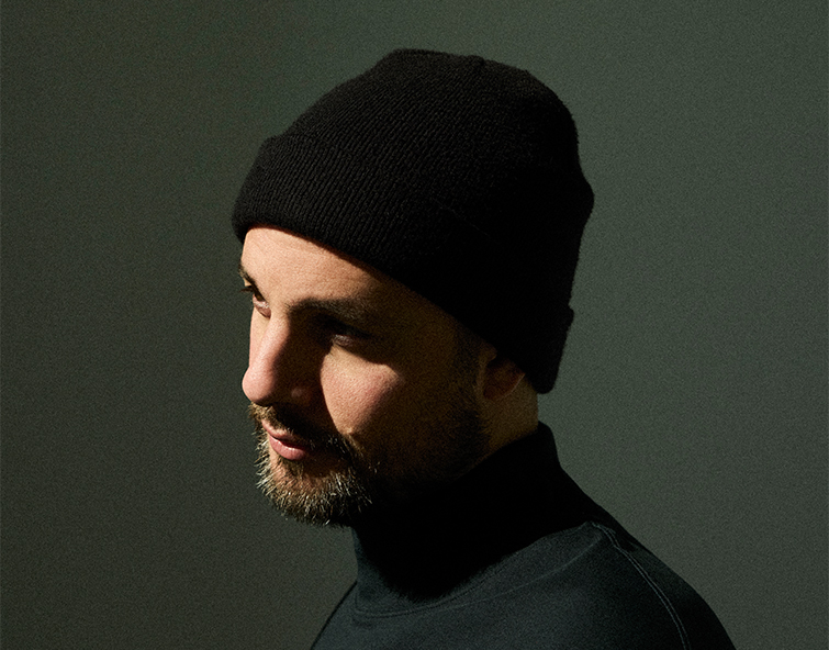 A moody portrait of a bearded Shai Maestro wearing a black beanie and dark turtleneck, lit from the side against a dark green background.