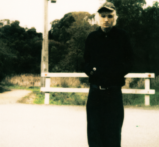 A full-length shot of Sweet Boy standing on an outdoor path with a grainy, vintage film aesthetic with blonde hair, neck tattoos, a dark baseball cap, a black jacket and black trousers.