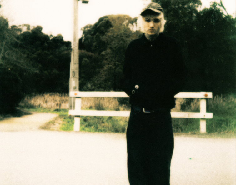 A full-length shot of Sweet Boy standing on an outdoor path with a grainy, vintage film aesthetic with blonde hair, neck tattoos, a dark baseball cap, a black jacket and black trousers.