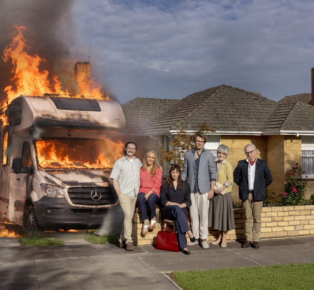 Six people posing for a family-style portrait in front of a brick house while an RV next to them is engulfed in large flames and thick smoke.