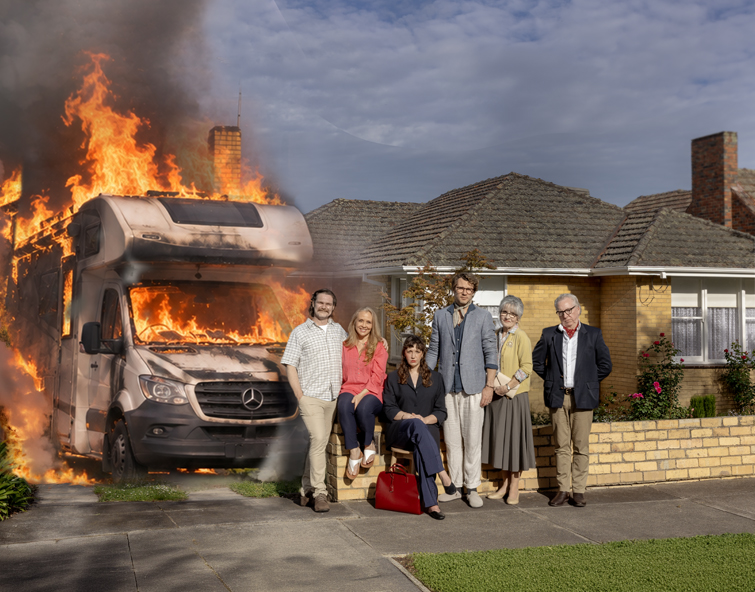 Six people posing for a family-style portrait in front of a brick house while an RV next to them is engulfed in large flames and thick smoke.